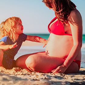 a young daughter placing her hand on her pregnant mothers belly while sitting on the beach